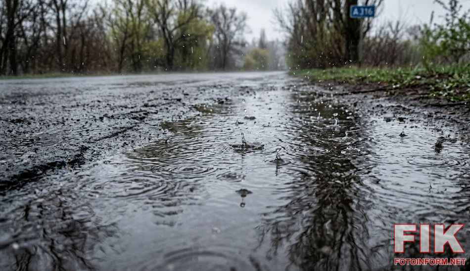 Крупний план калюжі на асфальті під час дощу в Україні. Видно бризки та кола на воді, похмурий пейзаж та дорожній знак на задньому плані. Ілюстрація до прогнозу погоди про опади.
